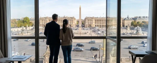 Un couple vu de dos regarde la Place de la Concorde depuis la fenêtre lumineuse d'une chambre d'hôtel parisienne, lumière matinale naturelle